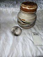 Overall view of pottery jar with lid and small round stone on white fabric background