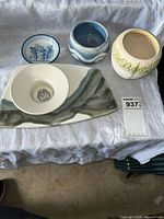 Four pottery and ceramic pieces on a white cloth, showing blue and white flower pot, small floral pot, white ceramic holder with metal spikes on a tray, and green-gray tray.