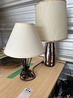 Two table lamps displayed on a wooden surface, one with painted pottery base and tall drum shade, the other with metal base and smaller conical shade.