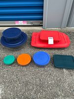 Photo of assorted Pyrex bowls and dishes with colored plastic lids outdoors on pavement, showing large rectangular dish with red lid, large round bowls with blue lids, and smaller round dishes with lids in varied colors.