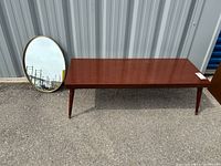 Overall view of the coffee table and oval mirror leaning against a metal wall, items placed on asphalt. Coffee table has a polished wooden surface with four tapered legs, the oval mirror has a brass frame.