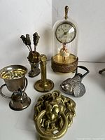 Overall lot showing vintage musical clock under glass dome, brass lion head door knocker, metal fish-shaped bottle opener, sterling silver candle snuffer, and brass holder with forks.