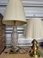 Two table lamps side by side: one a tall clear etched glass lamp with a tall beige pleated fabric shade, and the other a smaller brass lamp with a scalloped beige pleated shade. The glass lamp shade shows wear; the brass lamp base is noted as loose.