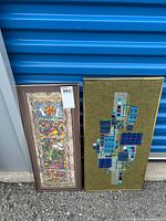Photo showing both framed wall art pieces against a blue metal storage unit door; left artwork is papyrus painted with abstract shapes, right artwork is mixed media with green textured background and geometric blue patterns.