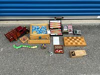 Overview photo of all wooden games and toys arranged on pavement including chess boards, backgammon set, wooden toy truck, playing cards, and puzzles.