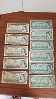 Top view of 16 Canadian banknotes including 1 Dollar and 2 Dollar bills arranged on a wooden surface, showing front sides with Queen Elizabeth II portrait.