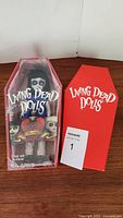 Living Dead Doll Rose in packaging next to red coffin-shaped box on wooden surface.