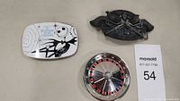 Three belt buckles side by side on surface: Nightmare Before Christmas, crossed pistols, and roulette wheel design.