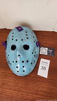 Front view of the light blue distressed hockey mask with multiple ventilation holes and purple adjustable straps.