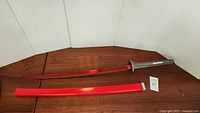 Full view of red-bladed katana-style sword with red scabbard on wooden surface.