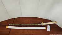 Full view of the cosplay sword laid beside its white sheath with yellow ends on a wooden surface.