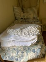 Twin bed made up with seaside themed bedding including a twin bedspread featuring seahorses and coral and layered with a white textured blanket with pom-pom trim at foot of bed. Two large striped pillows and an accent pillow with words.
