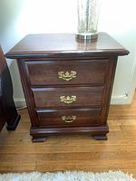Front view of one dark wood bedside/end table with three drawers and brass handles, showing full item detail and wood finish.