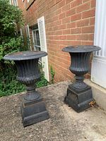 Two black cast iron urn planters side by side on concrete surface against brick wall