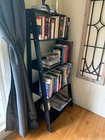 Black ladder style bookshelf fully assembled with books on shelves, positioned near a window with curtains.