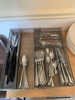 Set of cutlery arranged in metal mesh drawer organizer showing forks, spoons, knives and additional black handled knives.