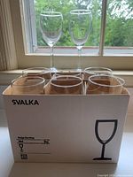 Photo of eight clear wine glasses placed inside the original IKEA SVALKA box with two additional glasses standing behind the box near a window.