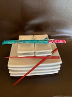 Stack of eight white square plates in two sizes with two sets of chopsticks on top, including one red pair and one pair in turquoise and floral wrap holders.