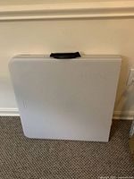 Closed folding table showing black handle and cream-colored plastic surface against wall.