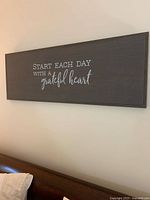 Front view of the gray wooden wall sign with white inspirational text mounted on a light colored wall above a bed.
