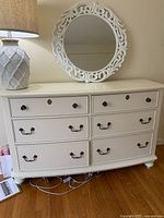 Full view of large white cream dresser with eight drawers and round ornate mirror on wall above it, placed on hardwood floor with a lamp on top