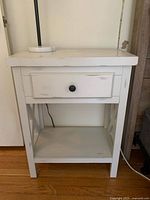 Front view of cream colored distressed end table with drawer and open shelf below on wooden floor.