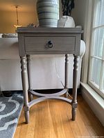Front view of gray end table showing the drawer with a black ring pull and turned legs on a hardwood floor near a sofa.