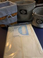 Photo showing the full lot with the cotton hanging dress bag in its package, two black and white zigzag patterned baskets, and two wicker baskets with blue gingham lining.