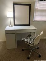 Photo showing white IKEA desk with drawer, white swivel chair with checkered cushion, silver desk lamp turned on, and black-framed bulletin board leaning against wall.