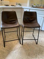 Two brown leather-like bar stools with black metal bases positioned on kitchen floor, showing front and seat shape.