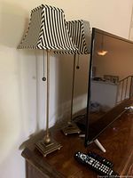 Pair of tall candlestick table lamps on wooden surface next to TV and remote. Lamps have gold-toned metal bases and black and white vertically striped shades.