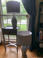 Pair of modern glass lamps with white drum shades in front of a window, one lamp on a table and one on floor.