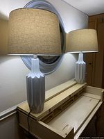 Front view of the pair of white porcelain lamps with beige textured drum shades on a cream-colored table underneath a large round wall clock.