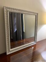Square white framed mirror leaning against a wall on wooden surface showcasing detailed carved frame and clear reflective glass.