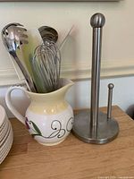 Photo showing ceramic pitcher with painted flowers holding cooking utensils next to stainless steel paper towel holder on wooden surface.