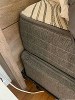Close-up view of mattress and boxspring corner showing gray fabric upholstery and mattress thickness.