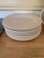Stack of 11 cream-colored dinner plates showing textured rope-like edge detail on a wooden surface.