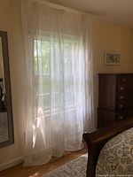 Sheer white curtain panel hanging in a bedroom window, showing delicate embroidered design and semi-transparency.