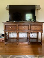Front view showing wood buffet console table with three drawers, round metal knobs, and flat top with TV and lamps.