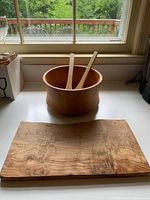 Williams Sonoma wooden salad bowl with two wooden tongs and live edge charcuterie board on countertop near window.