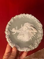 Front photo of small porcelain plate showing detailed lady with flowing hair and angel relief on light green background with floral border.