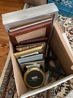 Box filled with assorted picture frames of various sizes and materials including wood, metal, and plastic. 4 black easels included.