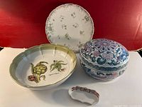 Front view of the four porcelain and ceramic decorative tableware pieces on red background, showing artichoke plate, floral-edged platter, lidded patterned bowl, and small decorative dish