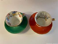Photo showing two Aynsley bone china teacup and saucer sets, one green and one orange, highlighting their floral interiors and gold trim.