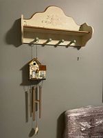 Image of the beige painted wooden wall shelf with floral designs, showing four green knobs and the hooked birdhouse wind chime hanging below.