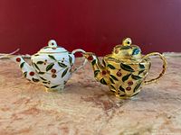 Photo showing front view of two small cloisonne metal teapot ornaments, one white with colored leaf design, one gold with colored leaf design