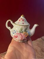 Miniature bone china tea pot held in hand showing floral design and gold trimming on lid and body.