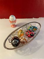Front angled view showing 5 hand-painted glass eggs inside elongated clear cut glass serving tray and one pink ceramic egg stand with a white-blue decorative egg on top.