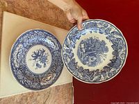 View of two blue and white porcelain plates side by side on a flat surface, one with floral motif and the other with rural scene.
