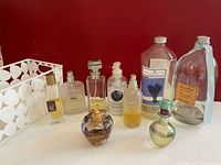 Photo showing 9 assorted perfume and fragrance bottles on a white shelf against a red background, displaying different sizes, shapes, and brands including a Lolita Lempicka apple-shaped bottle and a large bottle with lavender label.